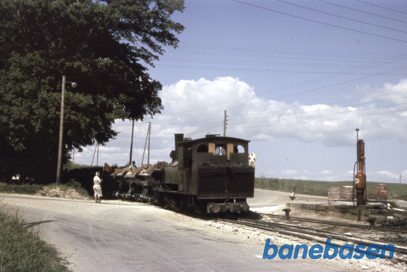 Vejskæring bevogtes af kvinde med rødt flag. T.h. tankanlæg for motorlokomotiver Vejskæring bevogtes af kvinde med rødt flag. T.h. tankanlæg for motorlokomotiver