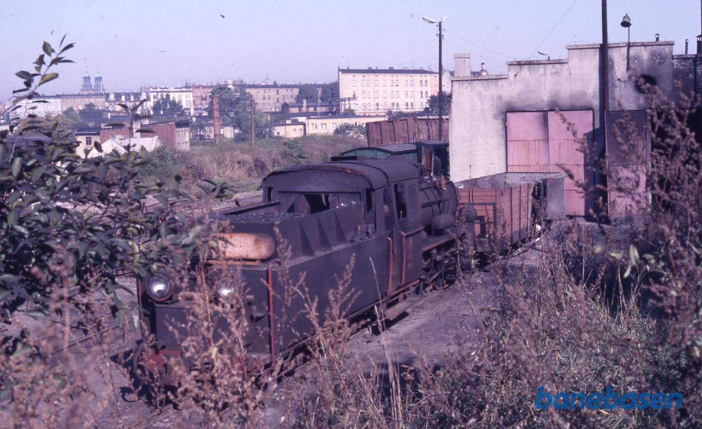 Damplokomotiv med godsvogn foran en remisebygning