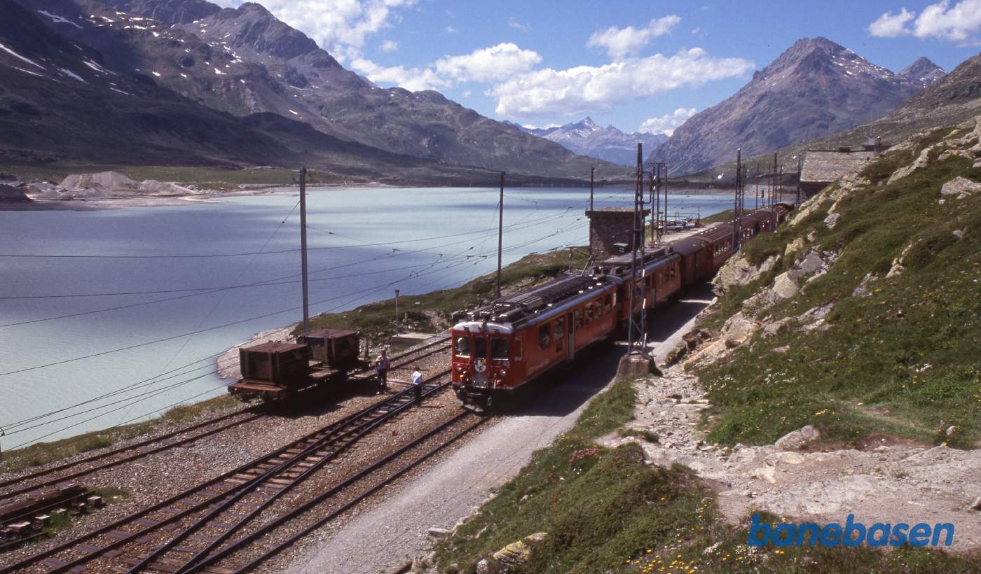 Stationen set skråt oppe fra med søen i baggrunden. Tog på vej mod Tirano venter på krydsning