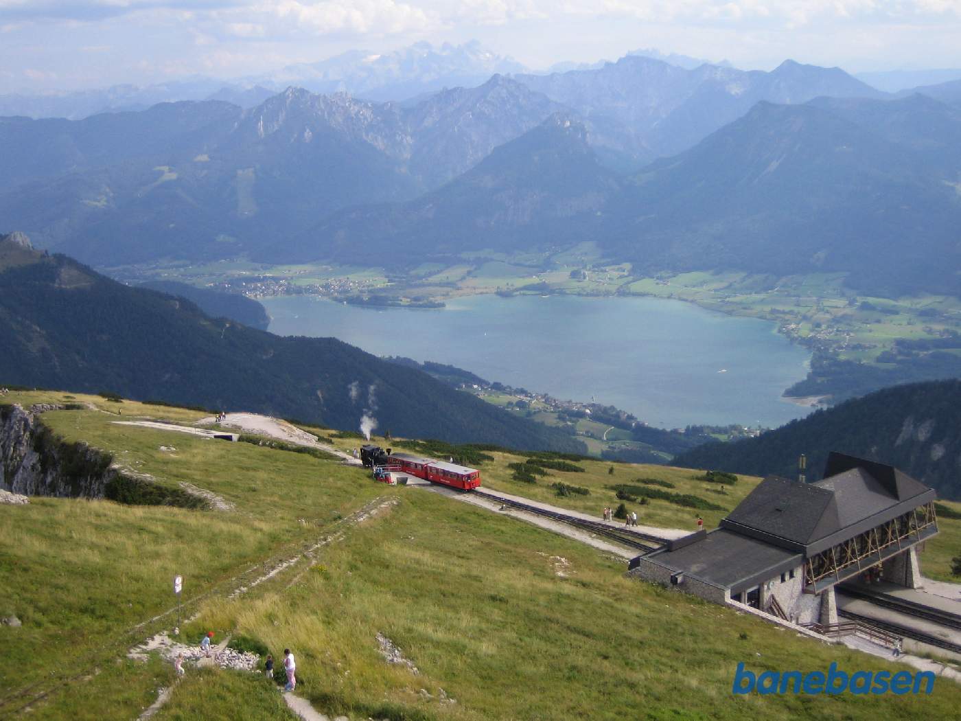 Et fyldt persontog på vej ned til byen ved søen. Lige foran toget ses det snedække (hvid beton) der ligner en tunnel