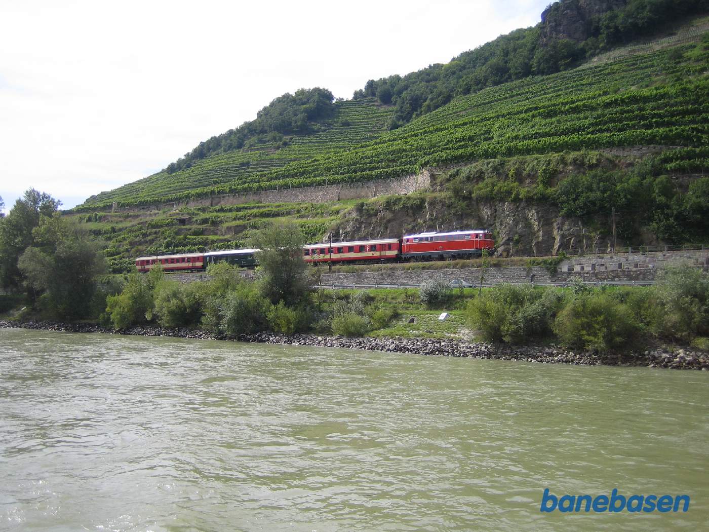 Under en sejltur på Donau, fik fotografen øje på et persontog på flodens nordlige bred, Toget kører i østlig retning. Bemærk vinmarkerne på skråningerne bag toget