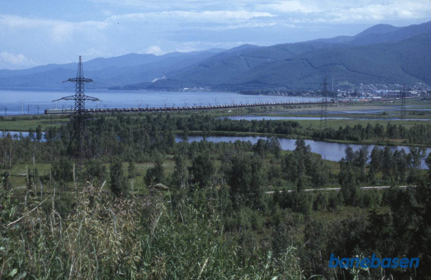 Et langt godstog på dæmningen langs søen. Nede foran kan jernbanen ud til industriområdet lige skimtes mellem træerne