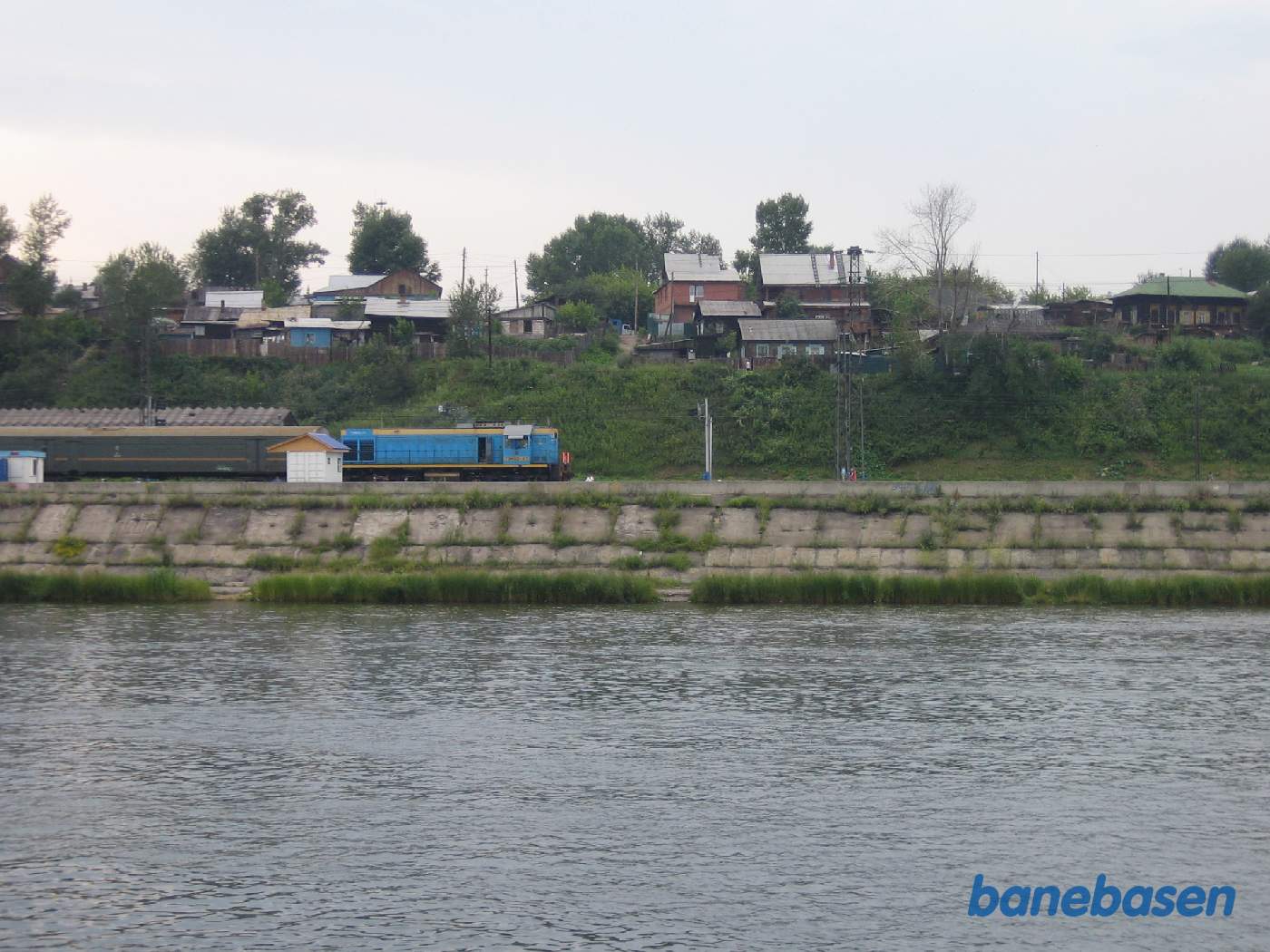 Et blåt diesellokomotiv med en grøn vogn, set fra flodens modsatte side