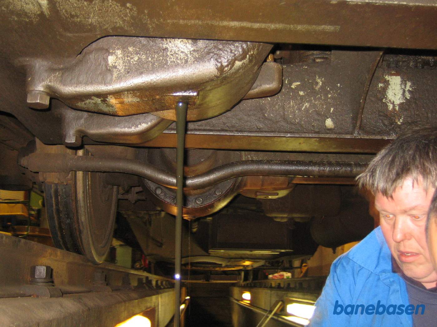 Olieskift på den forreste aksel på motorbogien, olien render ned i en spand og man kan se hvordan det drypper fra den anden aksel. Hjulakslen over hovedet på personen og den trækkende aksel over hovedet på fotografen