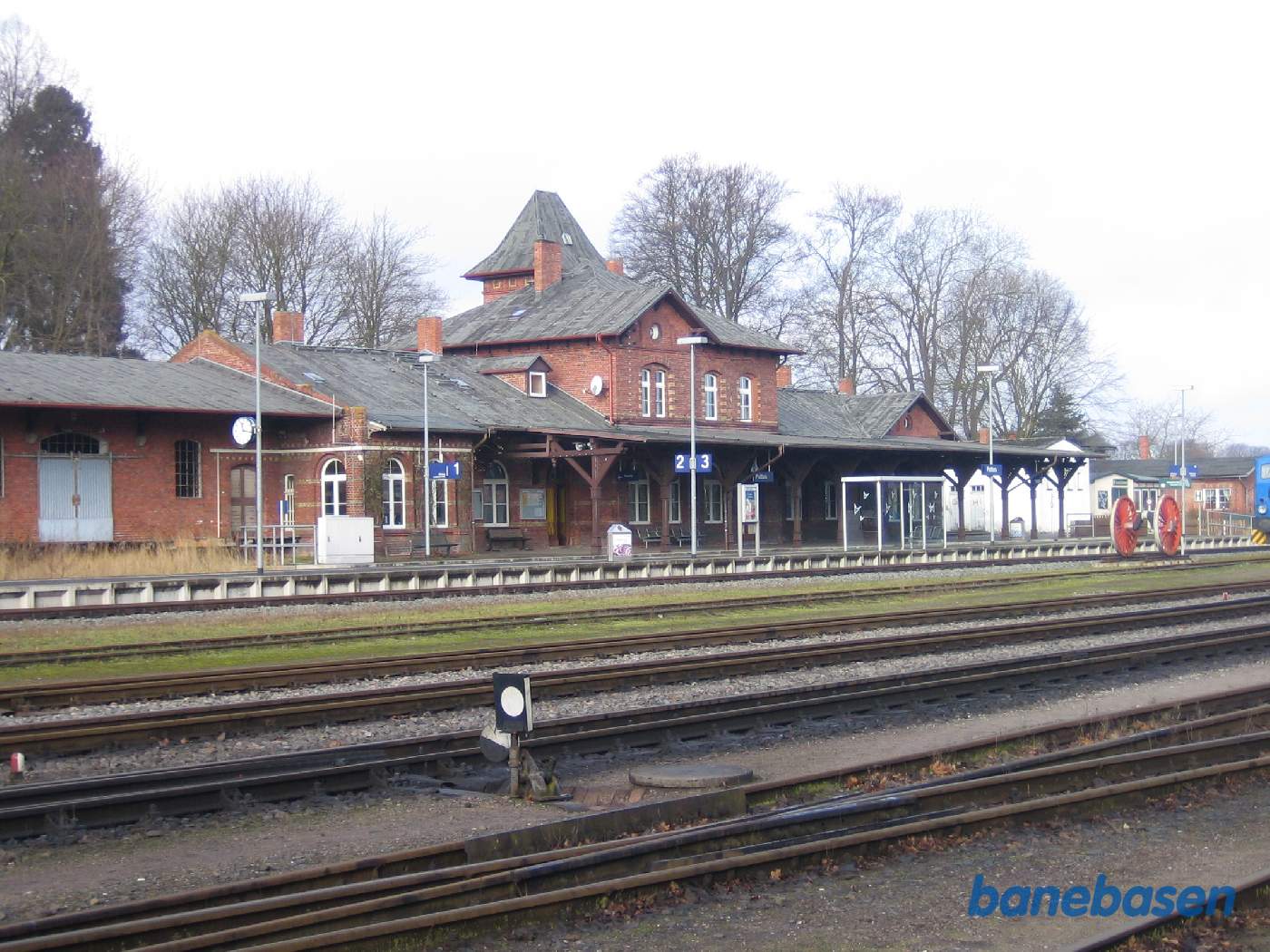Et vue ud over stationsområdet med stationsbygningen og et opstillet hjul fra et eksprestogsdamplokomotiv