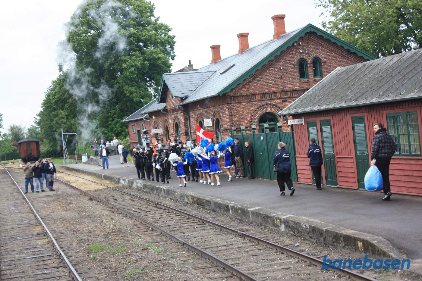 Musikkorpset marcherer foran stationsbygningen med dansepiger i følget Musikkorpset marcherer foran stationsbygningen med dansepiger i følget