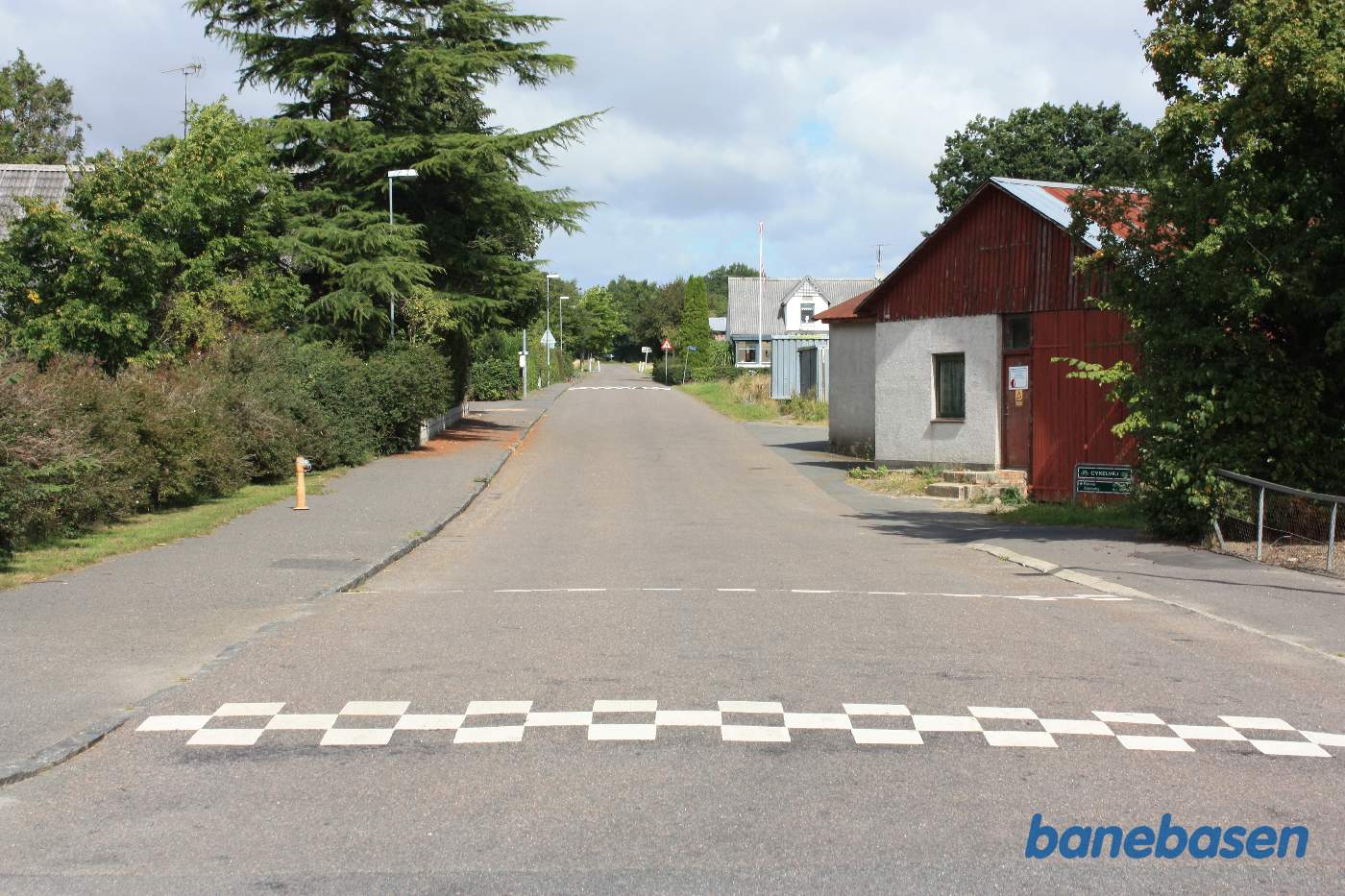 Vi står på Lobbæk Hovedgade. Cykelskiltet til højre markerer jernbaneoverkørslen. Stationen inde til venstre. Til højre fortsætter banen mod Nexø