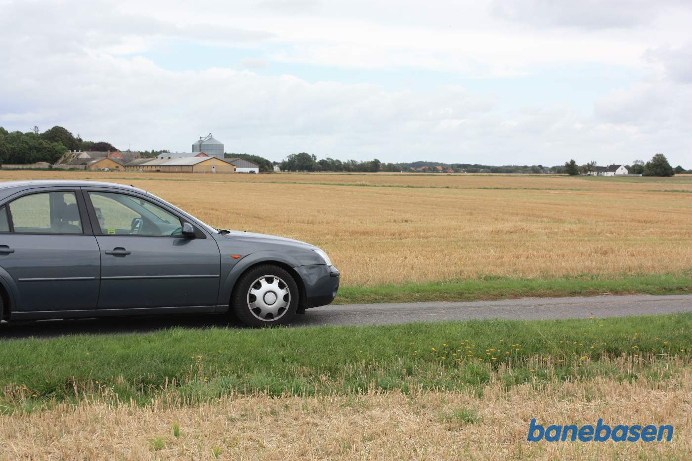 Vi er på Trassevejen og bilen markerer, hvor banen krydsede vejen og fortsatte over marken og gik lige til højre for den store gård over mod træerne. Vi ser i retning mod Nexø. Langemyregård trinbræt lå lige bag bilen.