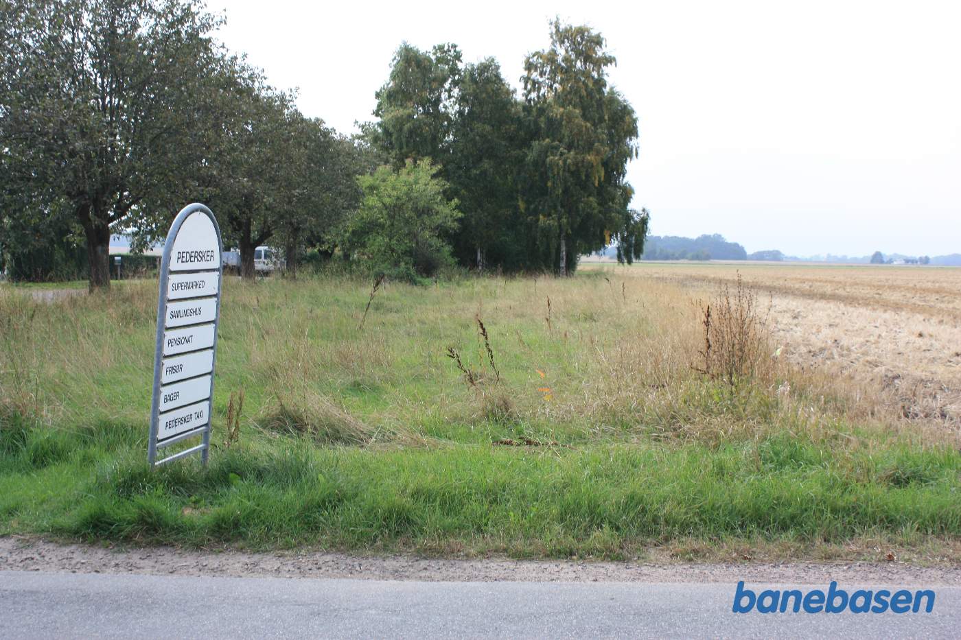 Vi er på Pedersker Hovedgade og kigger ind på stationsområdet. Vejen til stationen skimtes i billedets venstre side