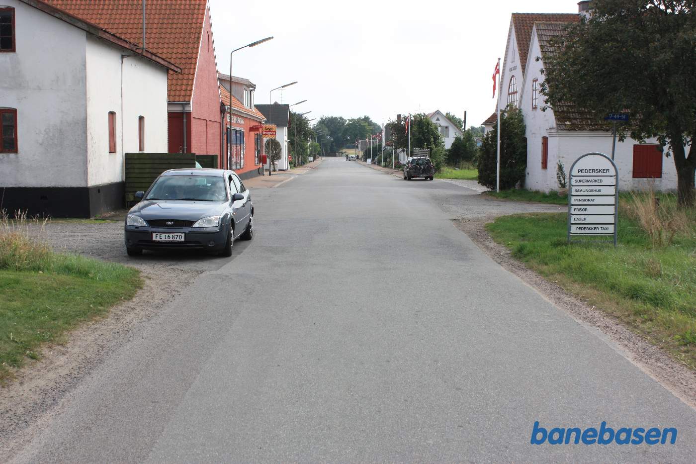 Vi er på Pedersker Hovedgade og bilen markerer hvor banen krydsede vejen, Stationen inde til højre og mod Nexø til venstre ned af grusvejen