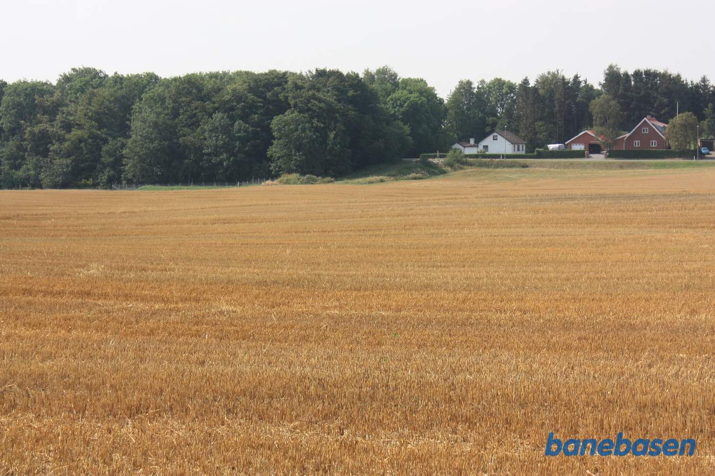 Vi står på marken på banetracéen og kigger mod Rønne. Banen gik på en høj banedæmning i en blød bue over marken og ind til venstre for det hvide hus