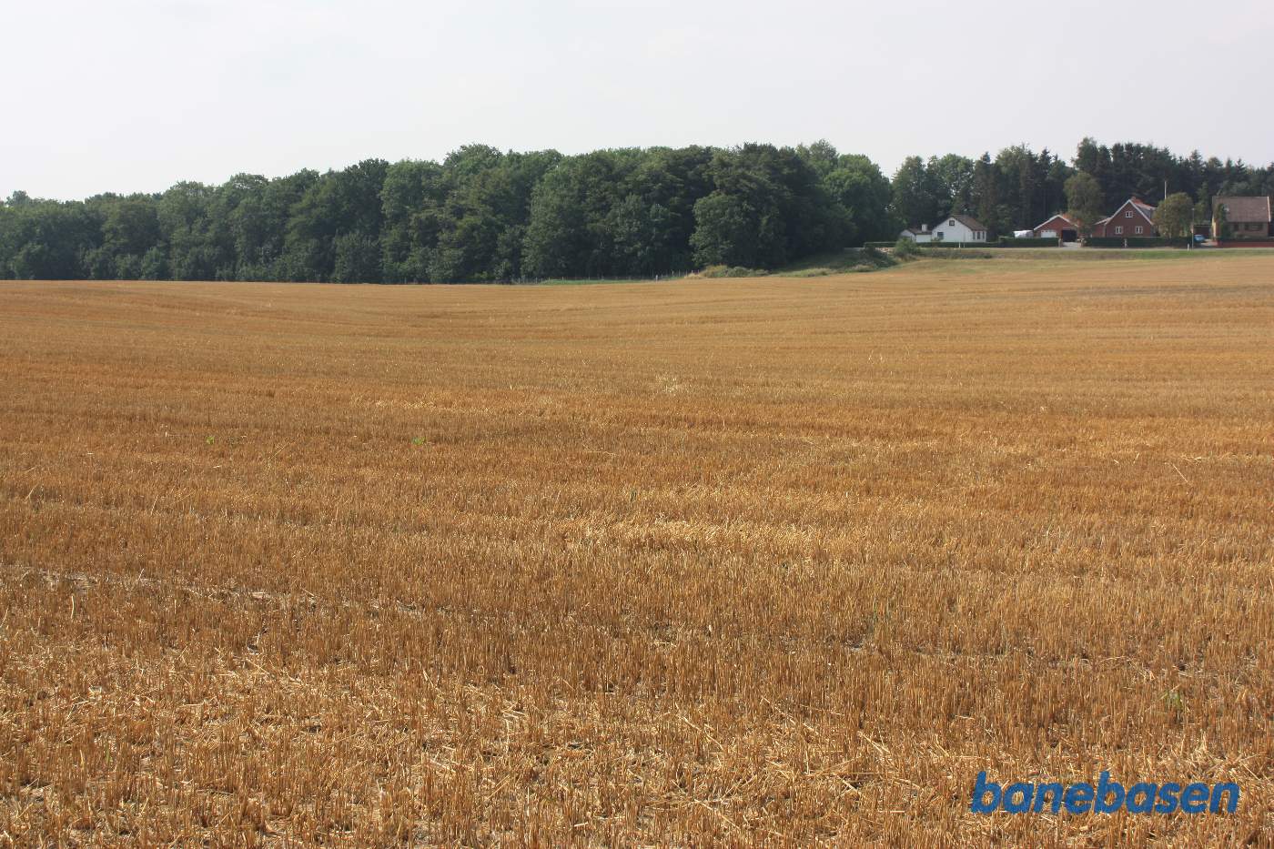 Vi står på marken på banetracéen og kigger mod Rønne. Banen gik på en høj banedæmning i en blød bue over marken og ind til venstre for det hvide hus Vi står på marken på banetracéen og kigger mod Rønne. Banen gik på en høj banedæmning i en blød bue over marken og ind til venstre for det hvide hus