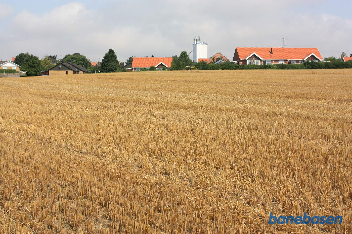 Vi står på marken på banetracéen og kigger mod Sandvig. Banen gik i en blød bue over marken og ind hvor den hvide tagvinkel på huset med rødt tag ligger midt i billedet