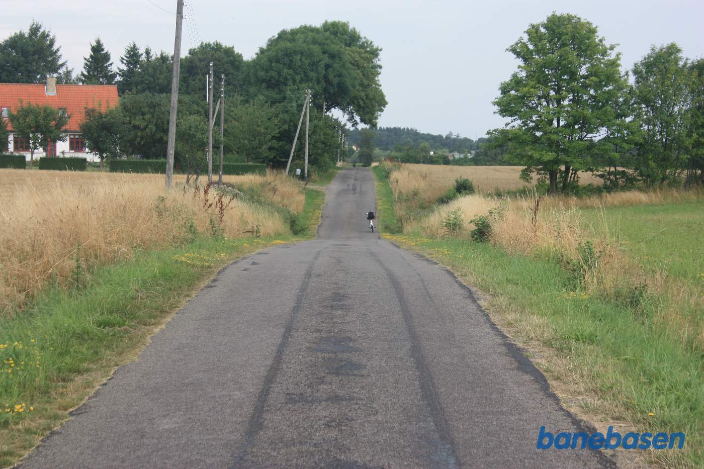 Vi står på Savmøllevej, hvor cyklen markerer overkørslen. Som det tydeligt ses, ligger banen forholdsvis lavt lige her, hvorfor vejen er sænket. Aakirkeby til venstre og Gudhjem til højre