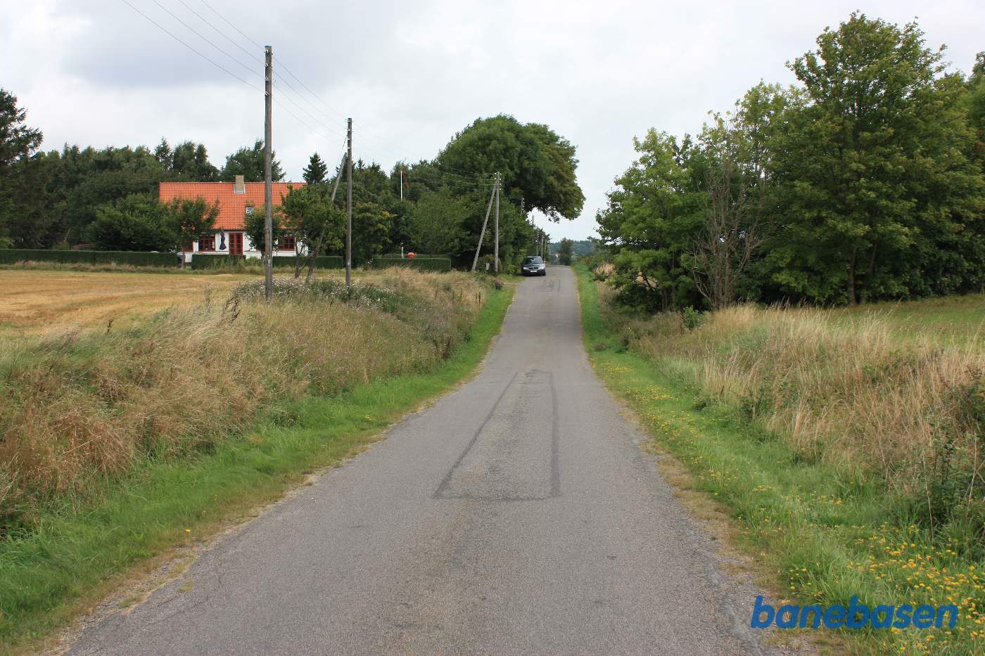 Vi står på Savmøllevej, hvor overkørslen lå nede i bunden af udgravningen. Som det tydeligt ses, ligger banen forholdsvis lavt lige her, hvorfor vejen er sænket. Aakirkeby til venstre og Gudhjem til højre