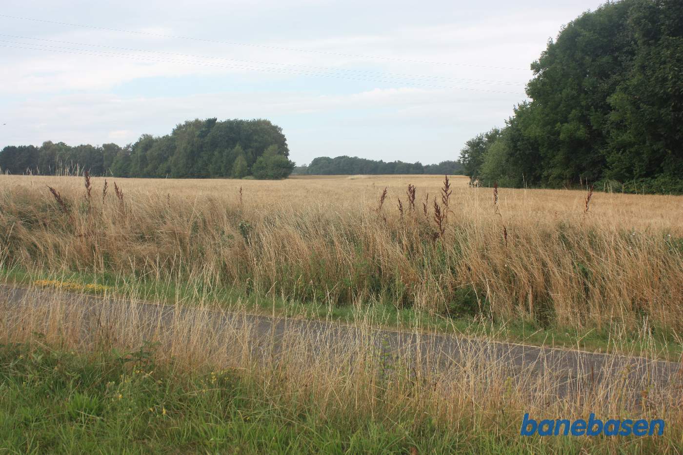 Vi står på Savmøllevej og kigger mod Aakirkeby. Banen gik inde mellem de 2 rækker af træer samt over det åbne markstykke Vi står på Savmøllevej og kigger mod Aakirkeby. Banen gik inde mellem de 2 rækker af træer samt over det åbne markstykke