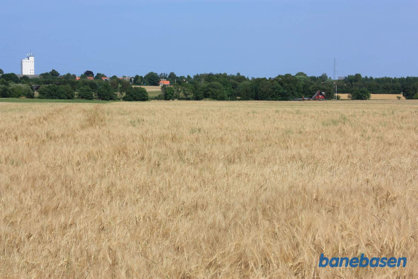 Vi står ude bag gården Fredensly og kigger mod Gudhjem. Der er ikke meget tilbage af banens tracé, men ser man godt efter, ses rasterne af banedæmningen ved skovkanten, lige midt i billedet