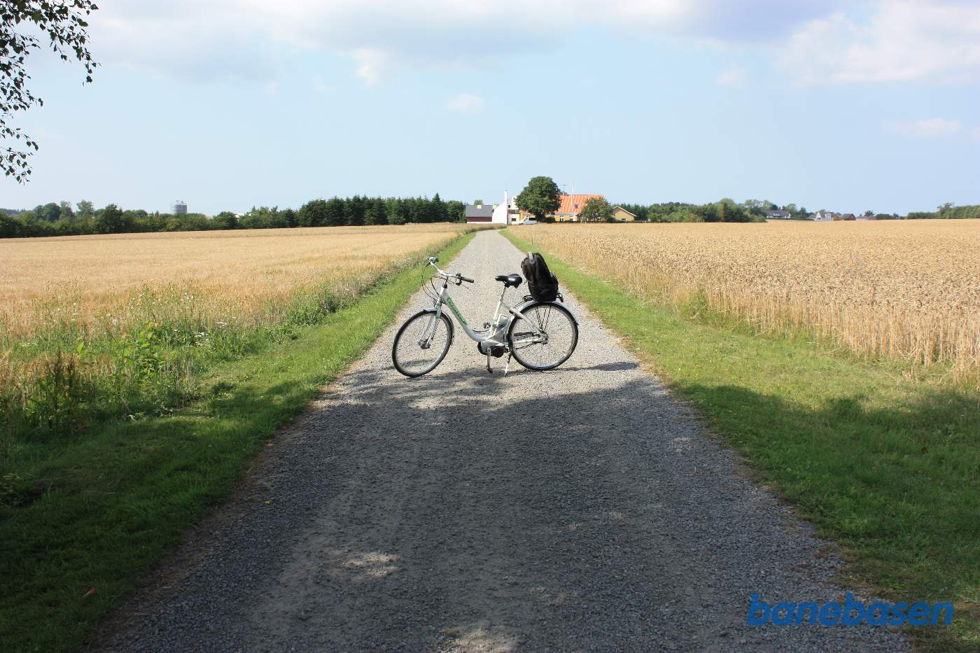 Vi står på en grusvej op til en stor gård. Cyklen markerer, hvor banen krydsede vejen. Aakirkeby til venstre og Gudhjem til højre Vi står på en grusvej op til en stor gård. Cyklen markerer, hvor banen krydsede vejen. Aakirkeby til venstre og Gudhjem til højre