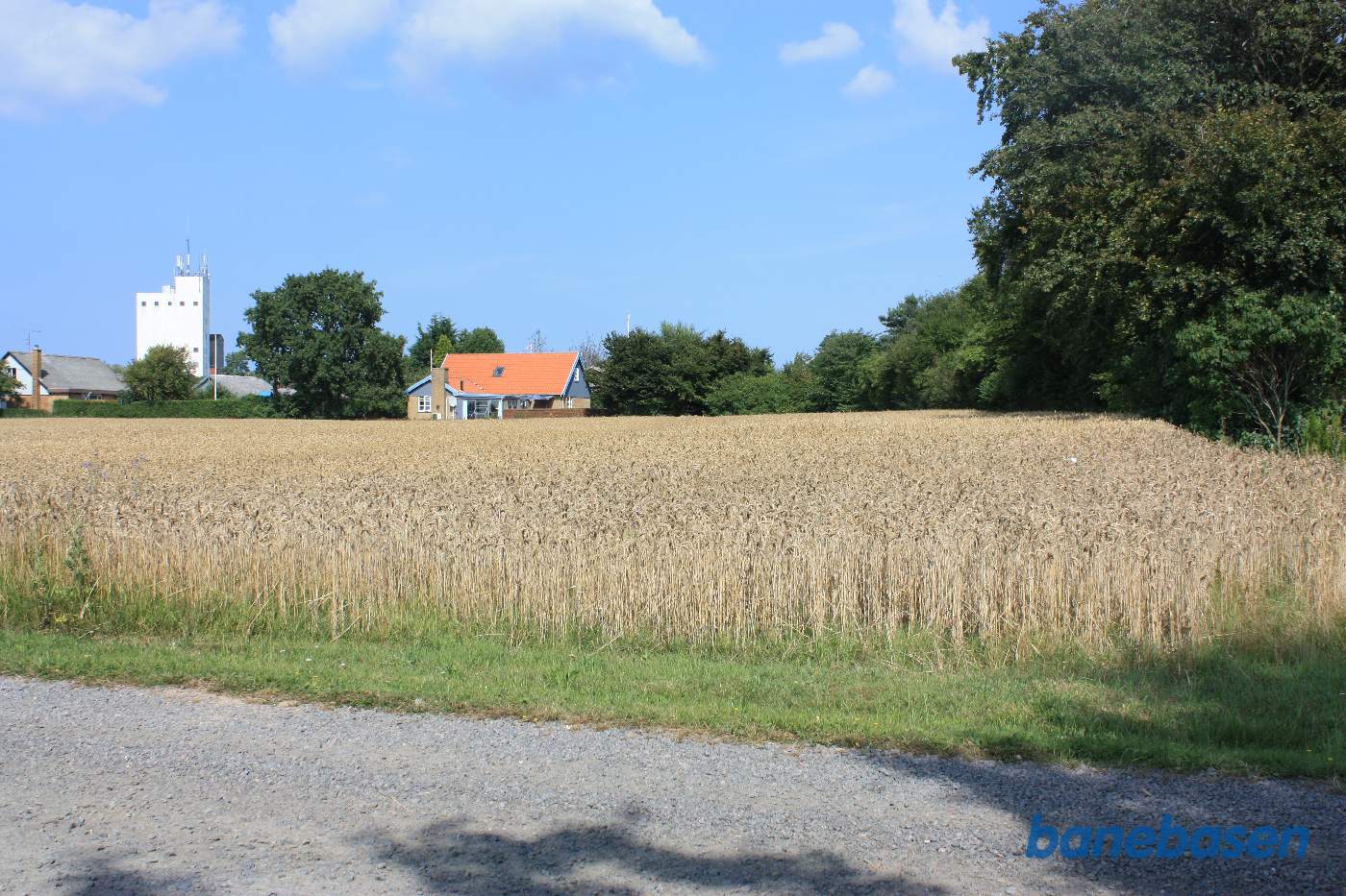 Vi står på en grusvej op til en stor gård. Vi kigger mod Gudhjem. Banen løb i en venstrekurve langs rækken af træer til højre, ind gennem huset med det røde tag og videre ind gennem det nye villakvarter til venstre