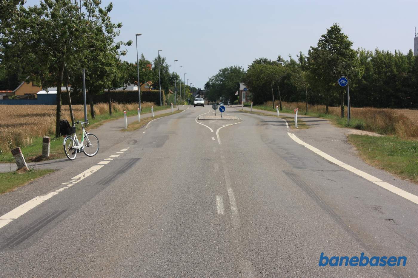 Vi står på Godthåbsvej, hvor banen krydsede vejen. På grund af den spidse vinkel, slog vejen et zigzag sving, så vejen kom vinkelret på banen. Cyklen markerer, hvor banen gik. Gudhjem til venstre og Aakirkeby skråt fremad. Stationsbygningen ligger nogenlunde ud for bilen til højre