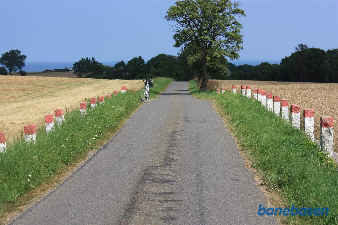 Vi står på Kjeldsebyvej. Cyklen markerer hvor banen krydsede vejen. Banen lå højere end vejen, hvilket tydeligt ses på billedet. Østersøen ses i baggrunden. Gudhjem til venstre og Aakirkeby til højre