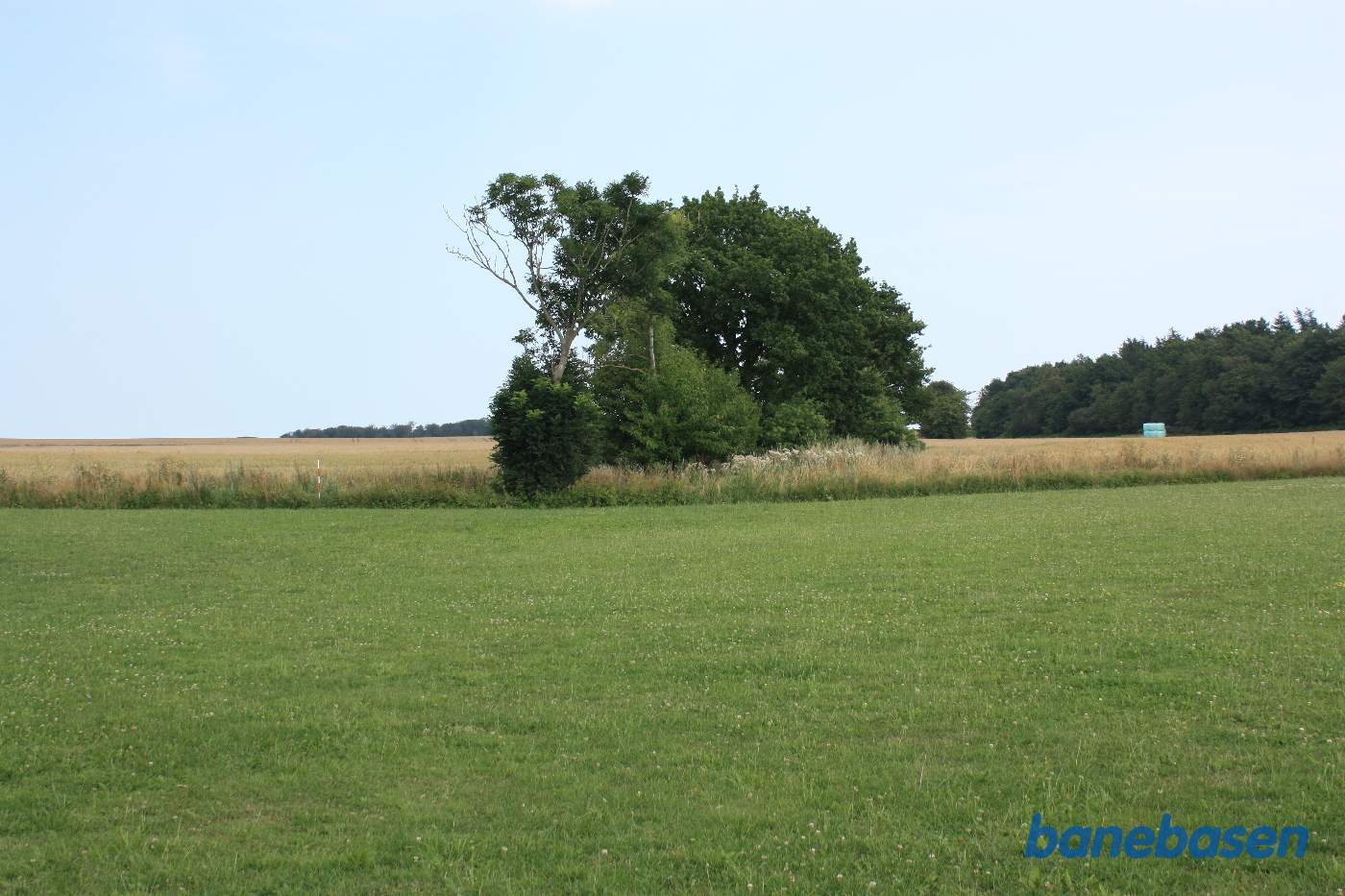 Vi står på en jordvej op til en gård og kigger i retning mod Aakirkeby. Banen gik inde i trærækken, over marken og videre igennem den næste trærække. Skellet for enden af græsplænen er en jordvej op til en anden gård