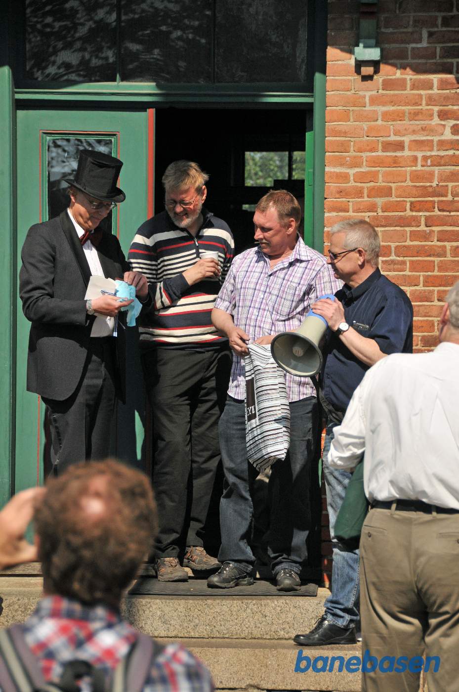 På trappen op til stationen, overrækkes de to fødselarer gaverne fra Jernbaneklubben, mens medlemmerne ser på På trappen op til stationen, overrækkes de to fødselarer gaverne fra Jernbaneklubben, mens medlemmerne ser på