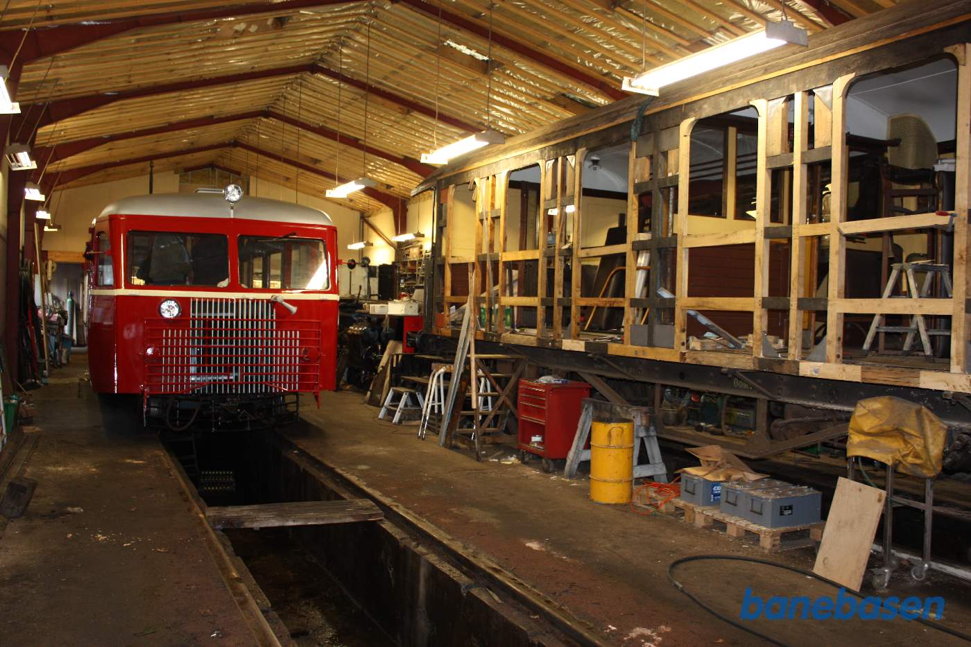En flot rød skinnebus i det ene værkstedsspor og en adskilt personvogn i det andet spor, hvor store dele af træskelettet udskiftes