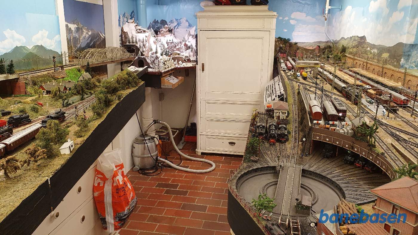 Et vue ud over hele den anden ende af modelbaneanlægget. Togene kører inde bag skabet, der er integreret i landskabet. Døren ind til kælderrummet ses til venstre, hvor de 2 løse "broer" muliggør kørsel rundt langs væggen Et vue ud over hele den anden ende af modelbaneanlægget. Togene kører inde bag skabet, der er integreret i landskabet. Døren ind til kælderrummet ses til venstre, hvor de 2 løse "broer" muliggør kørsel rundt langs væggen