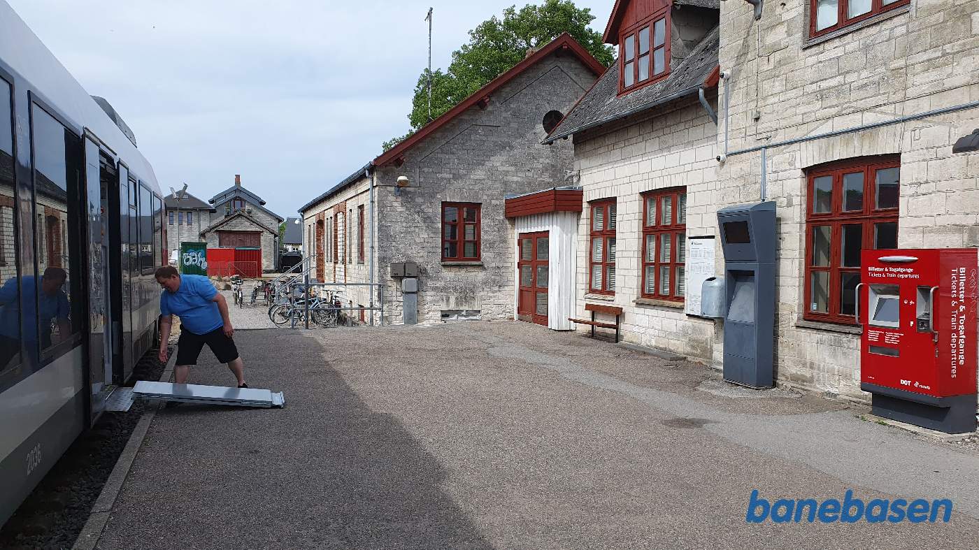 Lokomotivføreren folder kørestolsrampen ud, stationsbygningen inde til højre og remisen længere ude bag den røde container Lokomotivføreren folder kørestolsrampen ud, stationsbygningen inde til højre og remisen længere ude bag den røde container