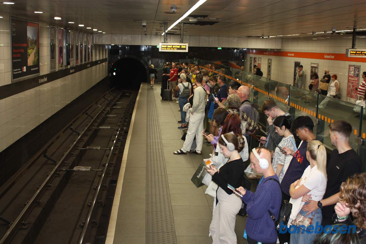 Et vue ud over perronen med mange passagerer, tog i modsat retning på den anden side af glasvæggen