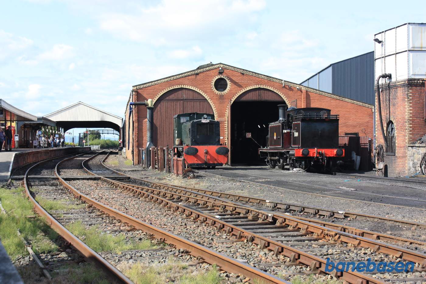 Et flot damplokomotiv står foran porten til remisen, perronsporet og stationsbygningen til venstre i billedet