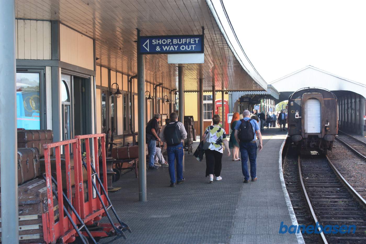 Personvognene holder ved perron og gæsterne begiver sig hen ad perronen, stationsbygningen til venstre